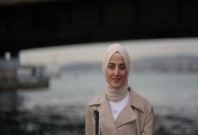 Görme engelli sosyolog ve kamu çalışanı Esra Durmuş, arka planda deniz ve iskele silüeti bulanık şekilde görünürken kameraya doğru hafif bir gülümsemeyle bakıyor.