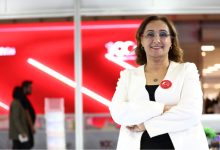 Modern bir iç mekânda, kırmızı arka plan önünde duran bir kadın kollarını bağlamış şekilde kameraya gülümsüyor. Üzerinde beyaz ceket ve gözlük var; yakasında rozet bulunuyor. Arka planda ise bulanık şekilde insanlar ve kurumsal bir ortam görülüyor.