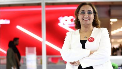 Modern bir iç mekânda, kırmızı arka plan önünde duran bir kadın kollarını bağlamış şekilde kameraya gülümsüyor. Üzerinde beyaz ceket ve gözlük var; yakasında rozet bulunuyor. Arka planda ise bulanık şekilde insanlar ve kurumsal bir ortam görülüyor.