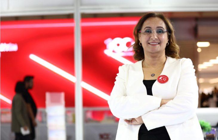 Modern bir iç mekânda, kırmızı arka plan önünde duran bir kadın kollarını bağlamış şekilde kameraya gülümsüyor. Üzerinde beyaz ceket ve gözlük var; yakasında rozet bulunuyor. Arka planda ise bulanık şekilde insanlar ve kurumsal bir ortam görülüyor.