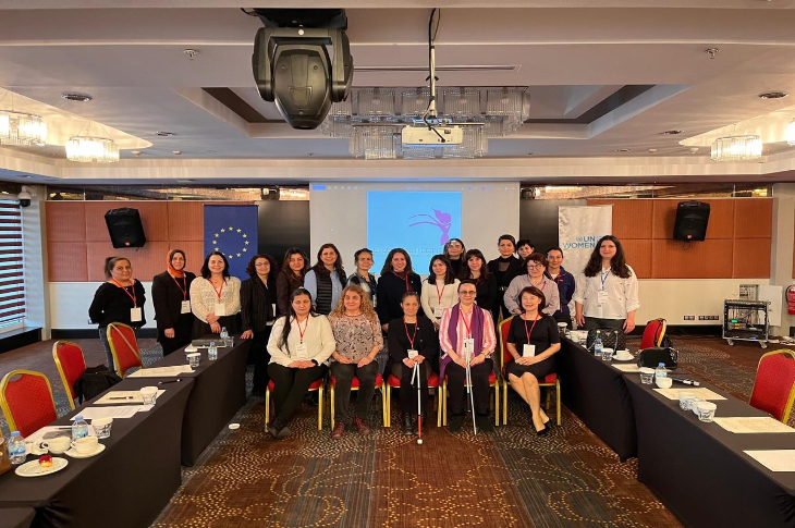 Bir toplantı salonunda, çoğunluğu kadınlardan oluşan kalabalık bir grup birlikte poz veriyor. Katılımcılar boyunlarında yaka kartlarıyla iki sıra halinde dizilmiş; bazıları otururken bazıları ayakta duruyor. Arka planda bir sunum perdesi ve etkinlik afişleri, yanlarda ise toplantı masaları ve sandalyeler görülüyor.