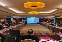 Geniş bir toplantı salonunda düzenlenmiş bir etkinlik görülüyor. Katılımcılar U şeklinde yerleştirilmiş masalarda oturuyor, ortada ise sunum yapan bir kişi ve projeksiyon ekranı var. Ekranda bilgi içeren slaytlar yer alırken, katılımcılar dikkatle sunumu dinliyor. Ortam resmi, düzenli ve eğitim/çalıştay havasında.