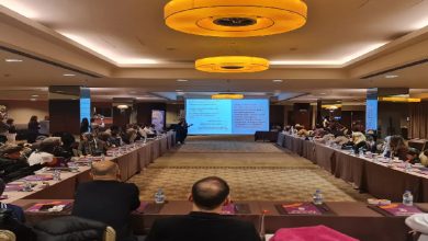 Geniş bir toplantı salonunda düzenlenmiş bir etkinlik görülüyor. Katılımcılar U şeklinde yerleştirilmiş masalarda oturuyor, ortada ise sunum yapan bir kişi ve projeksiyon ekranı var. Ekranda bilgi içeren slaytlar yer alırken, katılımcılar dikkatle sunumu dinliyor. Ortam resmi, düzenli ve eğitim/çalıştay havasında.