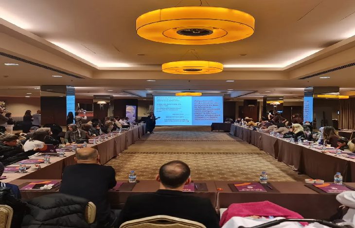 Geniş bir toplantı salonunda düzenlenmiş bir etkinlik görülüyor. Katılımcılar U şeklinde yerleştirilmiş masalarda oturuyor, ortada ise sunum yapan bir kişi ve projeksiyon ekranı var. Ekranda bilgi içeren slaytlar yer alırken, katılımcılar dikkatle sunumu dinliyor. Ortam resmi, düzenli ve eğitim/çalıştay havasında.