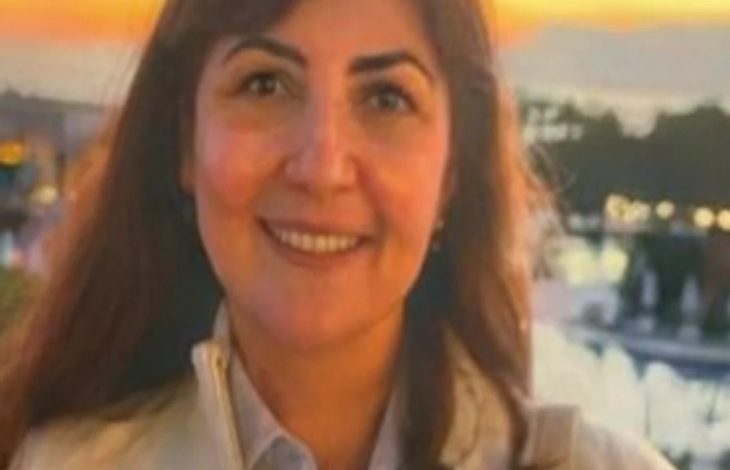 Gün batımı ışığında çekilmiş bir fotoğrafta, uzun koyu renk saçlı bir kadın kameraya gülümseyerek bakıyor. Açık renk bir ceket ve gömlek giymiş. Arka planda su kenarı, ağaçlar ve loş şehir ışıkları görülüyor; sıcak tonlu, sakin bir atmosfer hâkim.