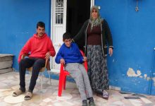 Görselde, mavi boyalı bir evin önünde üç kişi görülüyor. Ortada kırmızı plastik sandalyede oturan engelli bir genç, yanında sandalyede oturan başka bir genç ve arkalarında başörtülü bir kadın ayakta duruyor. Kadın, oturan gencin omzuna destek olur şekilde durarak birlikte kamera karşısında poz veriyor. Sahne, mütevazı bir evin avlusunda çekilmiş bir aile fotoğrafını andırıyor.