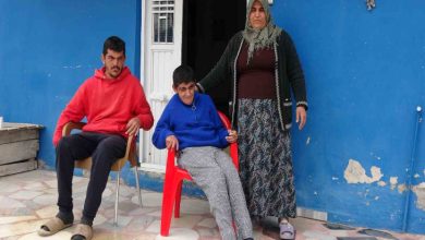 Görselde, mavi boyalı bir evin önünde üç kişi görülüyor. Ortada kırmızı plastik sandalyede oturan engelli bir genç, yanında sandalyede oturan başka bir genç ve arkalarında başörtülü bir kadın ayakta duruyor. Kadın, oturan gencin omzuna destek olur şekilde durarak birlikte kamera karşısında poz veriyor. Sahne, mütevazı bir evin avlusunda çekilmiş bir aile fotoğrafını andırıyor.