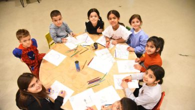 Bir sınıf ortamında küçük yaştaki çocuklar yuvarlak masa etrafında toplanmış, ellerinde boya kalemleriyle resim yapıyor. Masanın üzerinde dağınık halde kâğıtlar ve renkli kalemler bulunurken, çocuklar kameraya gülümseyerek poz veriyor.