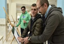 Bir üniversite koridorunda üç kişi kabartma bir tabloyu inceliyor. Ortadaki görme engelli birey, eserin yüzeyine dokunarak kabartma detayları hissederken yanında bulunan kişi ona rehberlik ediyor. Arka planda ise elinde beyaz baston bulunan başka bir görme engelli birey ve sergileme panoları görülüyor.