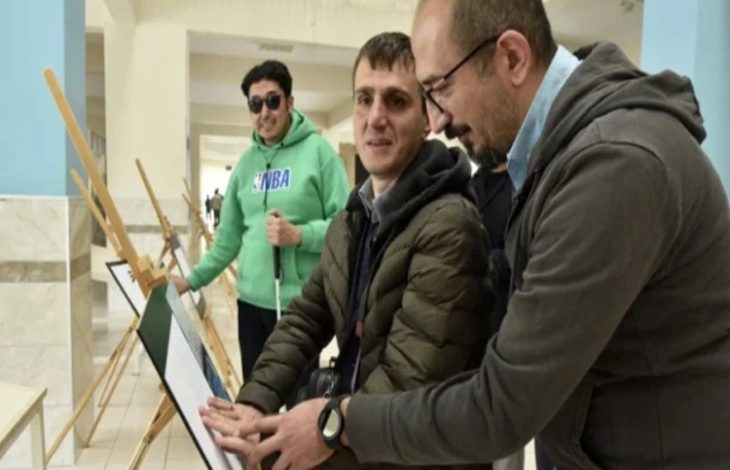 Bir üniversite koridorunda üç kişi kabartma bir tabloyu inceliyor. Ortadaki görme engelli birey, eserin yüzeyine dokunarak kabartma detayları hissederken yanında bulunan kişi ona rehberlik ediyor. Arka planda ise elinde beyaz baston bulunan başka bir görme engelli birey ve sergileme panoları görülüyor.