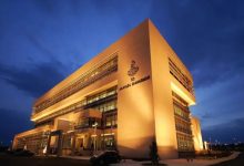 Fotoğrafta gece saatlerinde ışıklandırılmış modern bir kamu binası görülüyor. Binanın cephesinde Anayasa Mahkemesi yazısı ve amblemi yer alıyor. Geniş cam cepheli ve çok katlı yapı, resmi ve kurumsal mimarisiyle dikkat çekiyor.