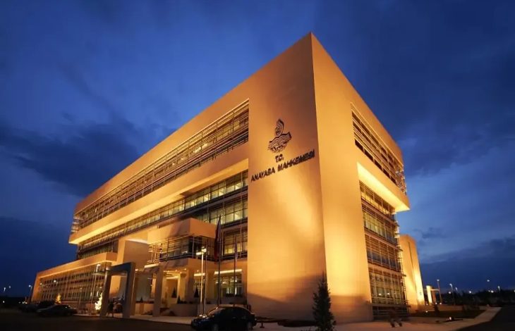 Fotoğrafta gece saatlerinde ışıklandırılmış modern bir kamu binası görülüyor. Binanın cephesinde Anayasa Mahkemesi yazısı ve amblemi yer alıyor. Geniş cam cepheli ve çok katlı yapı, resmi ve kurumsal mimarisiyle dikkat çekiyor.