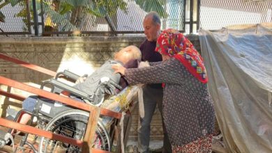 Görselde, tekerlekli sandalyede bulunan bir çocuğun taşınmasına yardımcı olan iki yetişkin görülüyor. Çocuk, eğimli ahşap bir rampada ilerletiliyor. Yanındaki kadın ve erkek, çocuğun güvenli bir şekilde hareket etmesi için destek veriyor. Arka planda duvar, bitki örtüsü ve gölgelik bir alan yer alıyor. Atmosfer, günlük yaşamda bakım ve destek gerektiren bir duruma işaret ediyor.