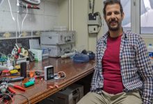 Elektronik ekipmanlarla dolu bir çalışma masasının yanında oturan biyomedikal yüksek mühendisi Oğuzhan Efe Sapmaz, kameraya gülümsüyor. Masanın üzerinde havya, kablolar, ölçüm cihazları ve çeşitli küçük elektronik parçalar bulunuyor.