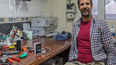 Elektronik ekipmanlarla dolu bir çalışma masasının yanında oturan biyomedikal yüksek mühendisi Oğuzhan Efe Sapmaz, kameraya gülümsüyor. Masanın üzerinde havya, kablolar, ölçüm cihazları ve çeşitli küçük elektronik parçalar bulunuyor.