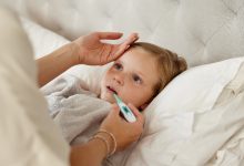 Yatakta yatan küçük bir çocuğun ateşi ölçülüyor. Bir yetişkin elini çocuğun alnına koyarken, diğer eliyle ağızdan ateş ölçer tutuyor. Çocuk halsiz ve hasta görünüyor; sahne ev ortamında bir hastalık durumunu yansıtıyor.