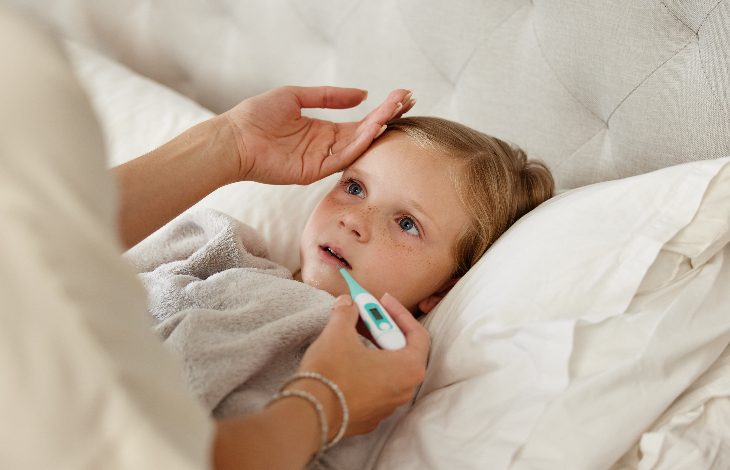 Yatakta yatan küçük bir çocuğun ateşi ölçülüyor. Bir yetişkin elini çocuğun alnına koyarken, diğer eliyle ağızdan ateş ölçer tutuyor. Çocuk halsiz ve hasta görünüyor; sahne ev ortamında bir hastalık durumunu yansıtıyor.