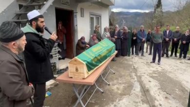 Bir imamın dua ettiği, etrafında insanların toplandığı ve yeşil örtüyle kaplı bir tabutun önünde gerçekleştirilen bir cenaze töreni görülüyor.