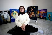 Fotoğrafta bir odada yerde oturan genç bir kadın görülüyor. Başörtülü olan kadın açık renk bir kazak giymiş ve kameraya doğru sakin bir ifadeyle bakıyor. Arka planda duvara dayalı şekilde sergilenen birkaç tablo bulunuyor. Tablolarda beyaz bir at, aslan figürü ve gün batımı manzarası gibi farklı resimler yer alıyor. Kadın, kendi yaptığı resimlerin önünde oturarak poz veriyor.