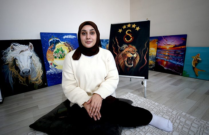 Fotoğrafta bir odada yerde oturan genç bir kadın görülüyor. Başörtülü olan kadın açık renk bir kazak giymiş ve kameraya doğru sakin bir ifadeyle bakıyor. Arka planda duvara dayalı şekilde sergilenen birkaç tablo bulunuyor. Tablolarda beyaz bir at, aslan figürü ve gün batımı manzarası gibi farklı resimler yer alıyor. Kadın, kendi yaptığı resimlerin önünde oturarak poz veriyor.