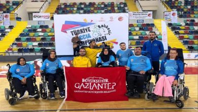 Bir spor salonunda, tekerlekli sandalyeli sporculardan oluşan Gaziantep Boccia Takımı, "Türkiye Boccia Şampiyonası" panosu önünde toplu poz veriyor. Sporcuların önünde "Büyükşehir Gaziantep" yazılı kırmızı bir bayrak açılmış durumda. Arka planda tribünler ve Türk bayrağı görülüyor.