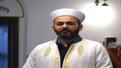 Geleneksel dini kıyafetler giymiş bir imam, kapalı bir mekânda kameraya doğru bakarak duruyor. Üzerinde beyaz cübbe ve sarık bulunuyor; arka planda ise cami içini andıran sade bir ortam görülüyor.
