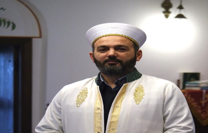 Geleneksel dini kıyafetler giymiş bir imam, kapalı bir mekânda kameraya doğru bakarak duruyor. Üzerinde beyaz cübbe ve sarık bulunuyor; arka planda ise cami içini andıran sade bir ortam görülüyor.