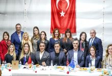 Fotoğrafta kapalı bir salonda düzenlenen bir iftar programında toplu halde poz veren bir grup görülüyor. Masanın önünde oturan ve arkada ayakta duran kadın ve erkeklerden oluşan kalabalık bir grup bulunuyor. Masanın üzerinde yemek tabakları, su şişeleri ve küçük bayraklar yer alıyor. Arka duvarda büyük bir Türk bayrağı asılı. Katılımcılar resmi kıyafetlerle kamera karşısında birlikte hatıra fotoğrafı çektiriyor.