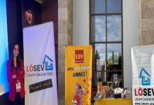 Fotoğrafta kapalı bir etkinlik alanında kurulan bir tanıtım standı görülüyor. Solda kırmızı ceketli bir kadın, LÖSEV (Lösemili Çocuklar Vakfı) logosunun yer aldığı bir afişin yanında duruyor. Ortada sarı renkte “LSV Dükkan – Ama seviyorum anne!” yazılı bir banner ve önünde çeşitli ürünlerin bulunduğu bir masa yer alıyor. Sağ tarafta ise yine LÖSEV logosunun bulunduğu başka bir afiş bulunuyor. Arka planda yüksek bir duvar, büyük bir pencere ve duvarda asılı bir portre görülüyor.