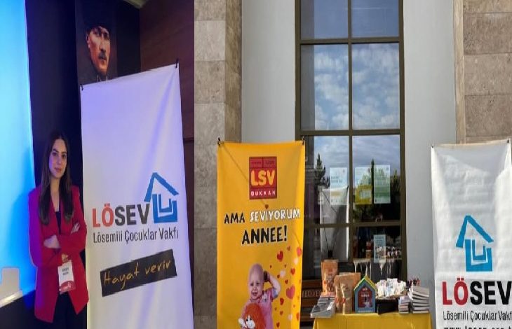 Fotoğrafta kapalı bir etkinlik alanında kurulan bir tanıtım standı görülüyor. Solda kırmızı ceketli bir kadın, LÖSEV (Lösemili Çocuklar Vakfı) logosunun yer aldığı bir afişin yanında duruyor. Ortada sarı renkte “LSV Dükkan – Ama seviyorum anne!” yazılı bir banner ve önünde çeşitli ürünlerin bulunduğu bir masa yer alıyor. Sağ tarafta ise yine LÖSEV logosunun bulunduğu başka bir afiş bulunuyor. Arka planda yüksek bir duvar, büyük bir pencere ve duvarda asılı bir portre görülüyor.