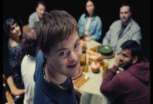 Bir yemek masasının etrafında toplanmış bir grup insan görülüyor. Ön planda, kameraya dönüp gülümseyen genç bir birey dikkat çekerken, masadaki diğer kişiler ona bakıyor. Ortam samimi bir aile ya da arkadaş buluşmasını andırıyor.