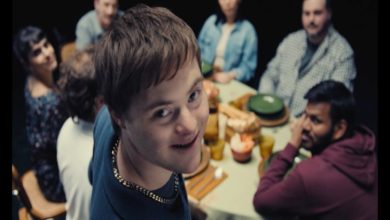 Bir yemek masasının etrafında toplanmış bir grup insan görülüyor. Ön planda, kameraya dönüp gülümseyen genç bir birey dikkat çekerken, masadaki diğer kişiler ona bakıyor. Ortam samimi bir aile ya da arkadaş buluşmasını andırıyor.