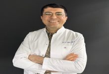 Fotoğrafta beyaz önlük giymiş bir diş hekimi görülüyor. Kollarını göğsünde birleştirmiş şekilde duruyor ve kameraya gülümseyerek poz vermiş. Yuvarlak çerçeveli gözlük takıyor. Arka plan koyu renk olduğu için önlük ve yüzü daha belirgin şekilde öne çıkıyor.