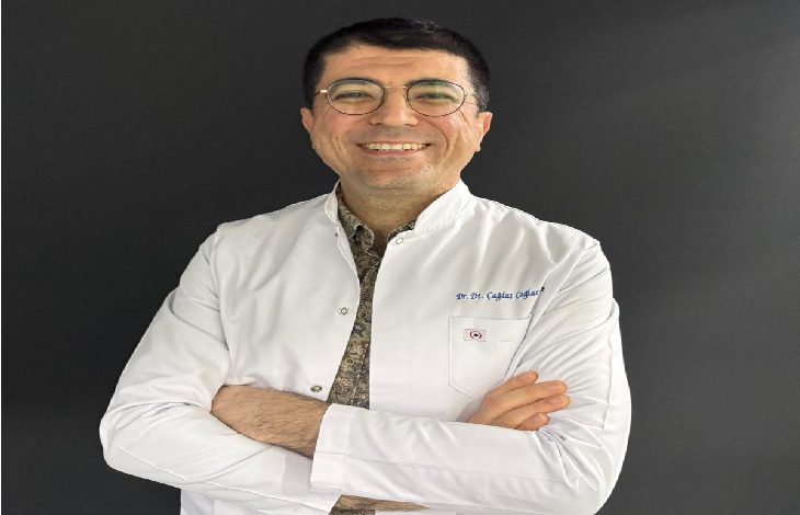 Fotoğrafta beyaz önlük giymiş bir diş hekimi görülüyor. Kollarını göğsünde birleştirmiş şekilde duruyor ve kameraya gülümseyerek poz vermiş. Yuvarlak çerçeveli gözlük takıyor. Arka plan koyu renk olduğu için önlük ve yüzü daha belirgin şekilde öne çıkıyor.