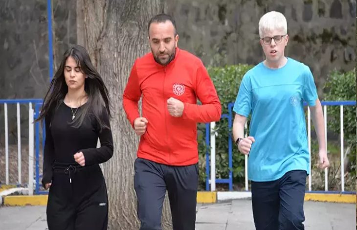 Açık alanda çekilmiş fotoğrafta üç kişi birlikte koşuyor. Ortada kırmızı eşofman üstü giyen bir yetişkin erkek, iki yanında ise spor kıyafetli gençler var. Soldaki siyah eşofmanlı genç kadın, sağdaki ise mavi tişörtlü genç erkek. Park benzeri bir ortamda, ağaç ve korkulukların bulunduğu bir alanda antrenman yaparken görüntülenmişler.