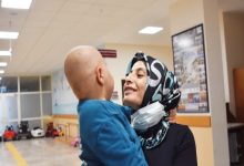 Bir hastane koridorunda başörtülü bir kadın, küçük bir çocuğu kucağında tutarak ona sevgiyle bakıyor. Çocuğun başı tıraşlı; bu da tedavi sürecine işaret ediyor. Kadının boynunda maske bulunurken ortamda umut ve şefkat hissi öne çıkıyor.