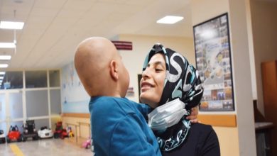 Bir hastane koridorunda başörtülü bir kadın, küçük bir çocuğu kucağında tutarak ona sevgiyle bakıyor. Çocuğun başı tıraşlı; bu da tedavi sürecine işaret ediyor. Kadının boynunda maske bulunurken ortamda umut ve şefkat hissi öne çıkıyor.