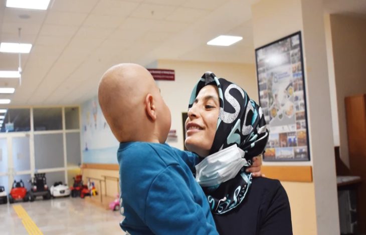Bir hastane koridorunda başörtülü bir kadın, küçük bir çocuğu kucağında tutarak ona sevgiyle bakıyor. Çocuğun başı tıraşlı; bu da tedavi sürecine işaret ediyor. Kadının boynunda maske bulunurken ortamda umut ve şefkat hissi öne çıkıyor.