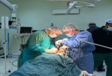 Bir ameliyathanede çekilen fotoğrafta, cerrahi kıyafet ve maskeler takan iki sağlık görevlisi ameliyat masasında yatan hastaya müdahale ediyor. Etraflarında ameliyat lambaları, tıbbi cihazlar ve monitörler bulunurken arka planda başka sağlık personeli de görülüyor.