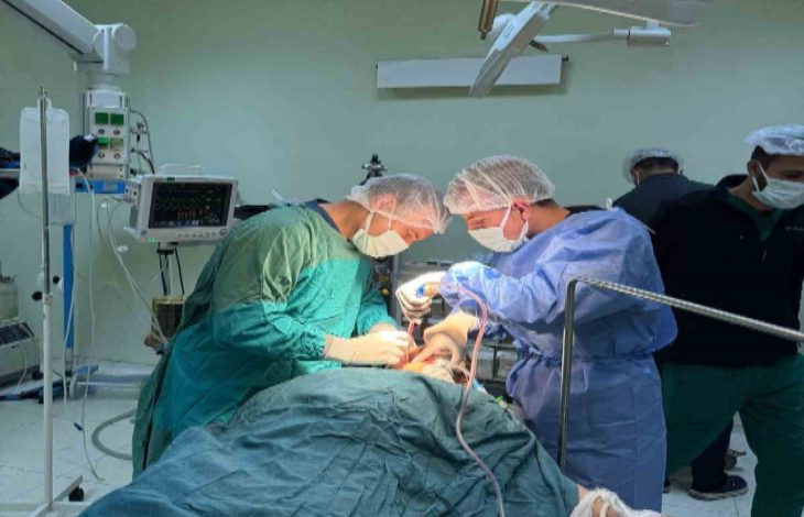 Bir ameliyathanede çekilen fotoğrafta, cerrahi kıyafet ve maskeler takan iki sağlık görevlisi ameliyat masasında yatan hastaya müdahale ediyor. Etraflarında ameliyat lambaları, tıbbi cihazlar ve monitörler bulunurken arka planda başka sağlık personeli de görülüyor.