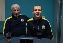 Fotoğrafta kapalı bir mekânda masada oturan iki erkek görülüyor. İkisi de lacivert ve sarı detaylı Fenerbahçe eşofman üstü giymiş. Soldaki yaşça daha büyük olan kişi beyaz saçlı ve bıyıklı; gülümseyerek yanındaki kişinin omzuna kolunu koymuş. Sağdaki kişi de kameraya doğru gülümseyerek oturuyor. Masanın üzerinde iki bardak çay ve bir telefon ya da küçük bir cihaz bulunuyor. Arka planda turkuaz renkli bir duvar ve dolap görülüyor.