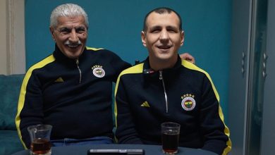 Fotoğrafta kapalı bir mekânda masada oturan iki erkek görülüyor. İkisi de lacivert ve sarı detaylı Fenerbahçe eşofman üstü giymiş. Soldaki yaşça daha büyük olan kişi beyaz saçlı ve bıyıklı; gülümseyerek yanındaki kişinin omzuna kolunu koymuş. Sağdaki kişi de kameraya doğru gülümseyerek oturuyor. Masanın üzerinde iki bardak çay ve bir telefon ya da küçük bir cihaz bulunuyor. Arka planda turkuaz renkli bir duvar ve dolap görülüyor.