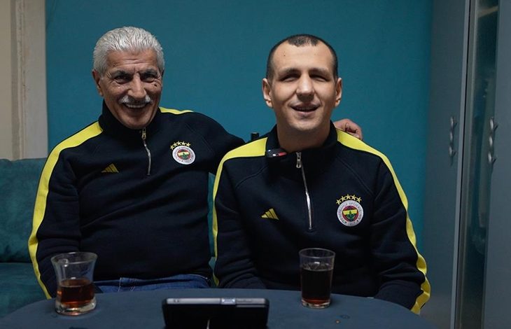 Fotoğrafta kapalı bir mekânda masada oturan iki erkek görülüyor. İkisi de lacivert ve sarı detaylı Fenerbahçe eşofman üstü giymiş. Soldaki yaşça daha büyük olan kişi beyaz saçlı ve bıyıklı; gülümseyerek yanındaki kişinin omzuna kolunu koymuş. Sağdaki kişi de kameraya doğru gülümseyerek oturuyor. Masanın üzerinde iki bardak çay ve bir telefon ya da küçük bir cihaz bulunuyor. Arka planda turkuaz renkli bir duvar ve dolap görülüyor.