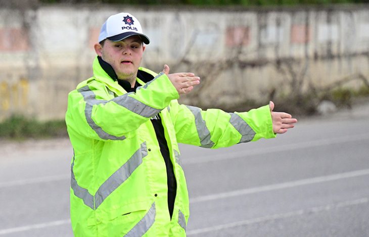 Neon sarı reflektörlü mont ve “polis” yazılı şapka giyen Down sendromlu Fırat Öztürk, yolda araçların yönünü elleriyle işaret ederek trafiği düzenliyor.