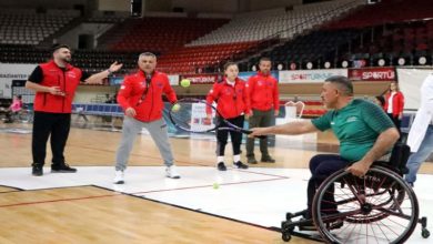 Bir spor salonunda, tekerlekli sandalyedeki bir adam raketle topa vururken, karşısında kırmızı ceketli birkaç kişi oyunu izleyip yönlendiriyor.