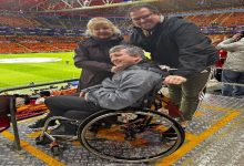 Fotoğrafta bir futbol stadyumunun tribününde tekerlekli sandalyede oturan bir taraftar görülüyor. Yanında bulunan iki kişiyle birlikte kameraya gülümseyerek poz veriyor. Arka planda ise sarı-kırmızı koltuklarıyla dolu büyük bir stadyum ve futbol sahası yer alıyor.