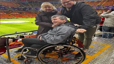 Fotoğrafta bir futbol stadyumunun tribününde tekerlekli sandalyede oturan bir taraftar görülüyor. Yanında bulunan iki kişiyle birlikte kameraya gülümseyerek poz veriyor. Arka planda ise sarı-kırmızı koltuklarıyla dolu büyük bir stadyum ve futbol sahası yer alıyor.