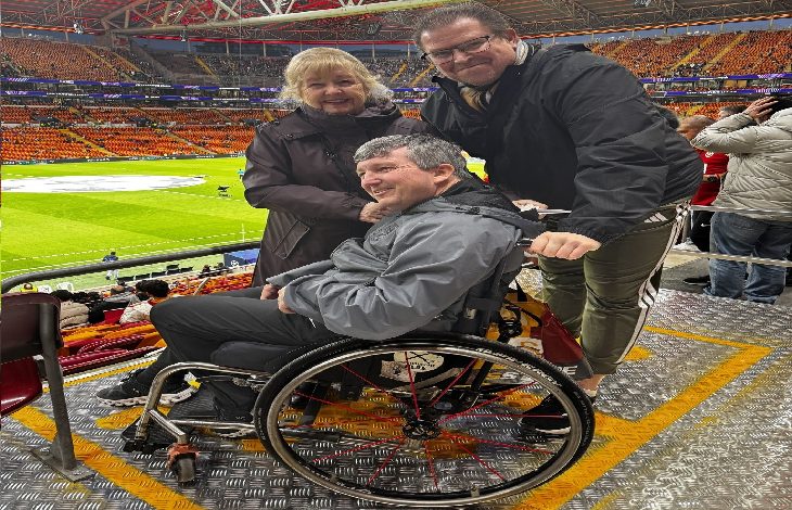 Fotoğrafta bir futbol stadyumunun tribününde tekerlekli sandalyede oturan bir taraftar görülüyor. Yanında bulunan iki kişiyle birlikte kameraya gülümseyerek poz veriyor. Arka planda ise sarı-kırmızı koltuklarıyla dolu büyük bir stadyum ve futbol sahası yer alıyor.