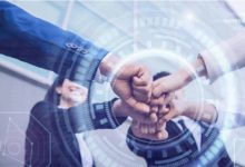 Mavi tonların hakim olduğu görselde, merkezde üst üste kenetlenmiş eller etrafında dijital halkalar ve teknoloji grafikleri yer alıyor. Arka planda ise iş birliğini simgeleyen, gülümseyen takım elbiseli profesyoneller görülüyor; genel tema birlik ve inovasyon.