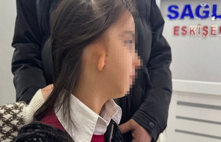 Görselde, yüzü gizlenmiş küçük bir kız çocuğu profilden görülüyor. Sağ kulak bölgesinde doğuştan gelişim farklılığı dikkat çekiyor. Yanında bir yetişkin destek amaçlı dururken, arka planda bir sağlık kurumuna ait tabela bulunuyor. Ortam, bir muayene ya da hastane sürecini çağrıştırıyor.